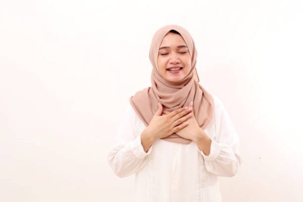 Portrait of relieved young asian muslim woman standing while holding chest and closing eyes